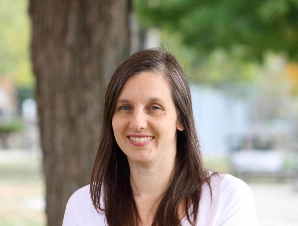 A woman with long brown hair and a white shirt smiles outdoors near a tree, with green leaves in the background.