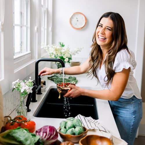 Woman smiling while rinsing strawberries at a kitchen sink with fresh vegetables on the counter.