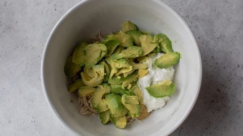 A white bowl with sliced avocado, shredded chicken, and a dollop of yogurt on a gray surface.