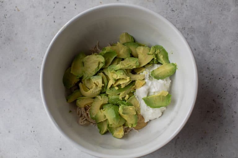 A white bowl with sliced avocado, shredded chicken, and a dollop of yogurt on a gray surface.