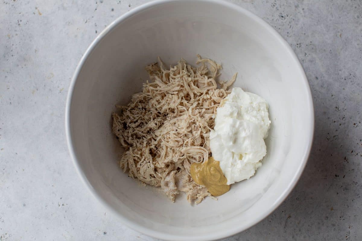 White bowl with shredded chicken, a dollop of Greek yogurt, mustard, and seasoning on a light gray surface.
