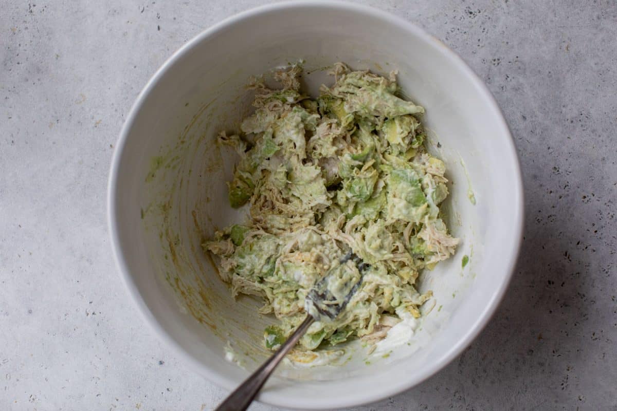 Shredded chicken, avocado, and mayonnaise mixture in a white bowl with a fork on a light gray surface.
