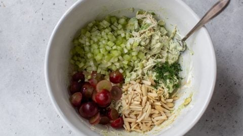A white bowl with celery, grapes, slivered almonds, dill, and creamy avocado chicken salad, with a spoon.
