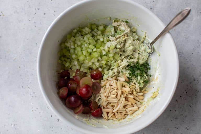 A white bowl with celery, grapes, slivered almonds, dill, and creamy avocado chicken salad, with a spoon.