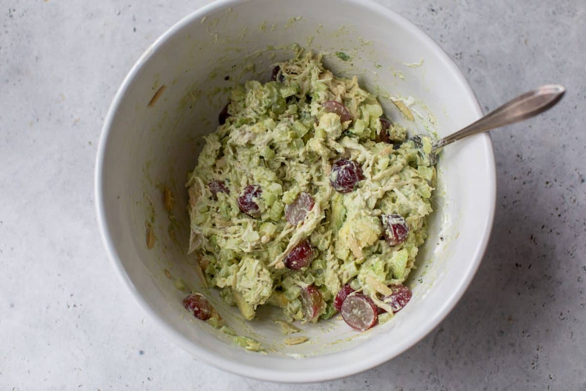 A bowl of chicken salad with avocado, celery, and red grapes, mixed with a spoon on a gray surface.