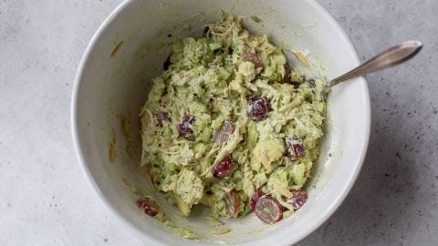A bowl of chicken salad with avocado, celery, and red grapes, mixed with a spoon on a gray surface.