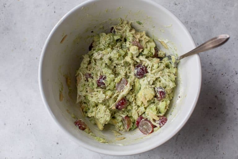 A bowl of chicken salad with avocado, celery, and red grapes, mixed with a spoon on a gray surface.
