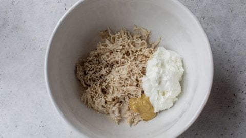 White bowl with shredded chicken, a dollop of Greek yogurt, mustard, and seasoning on a light gray surface.