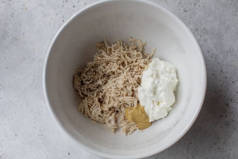 White bowl with shredded chicken, a dollop of Greek yogurt, mustard, and seasoning on a light gray surface.