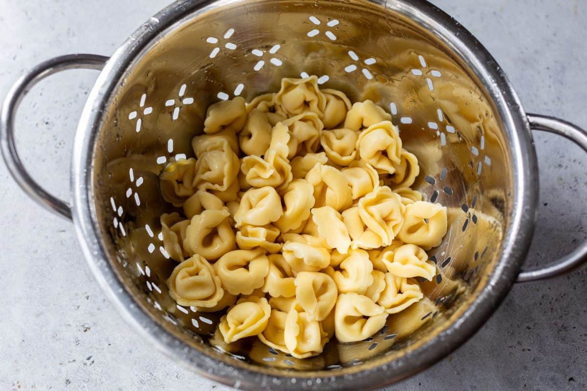 Cooked tortellini pasta draining in a metal colander on a light gray surface.