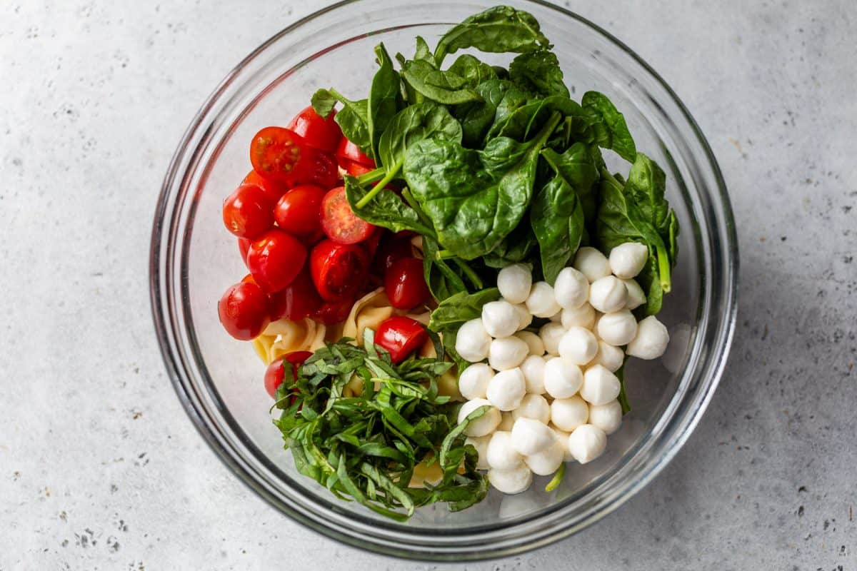 Glass bowl with cherry tomatoes, spinach, mozzarella balls, pasta, and fresh basil on a light surface.