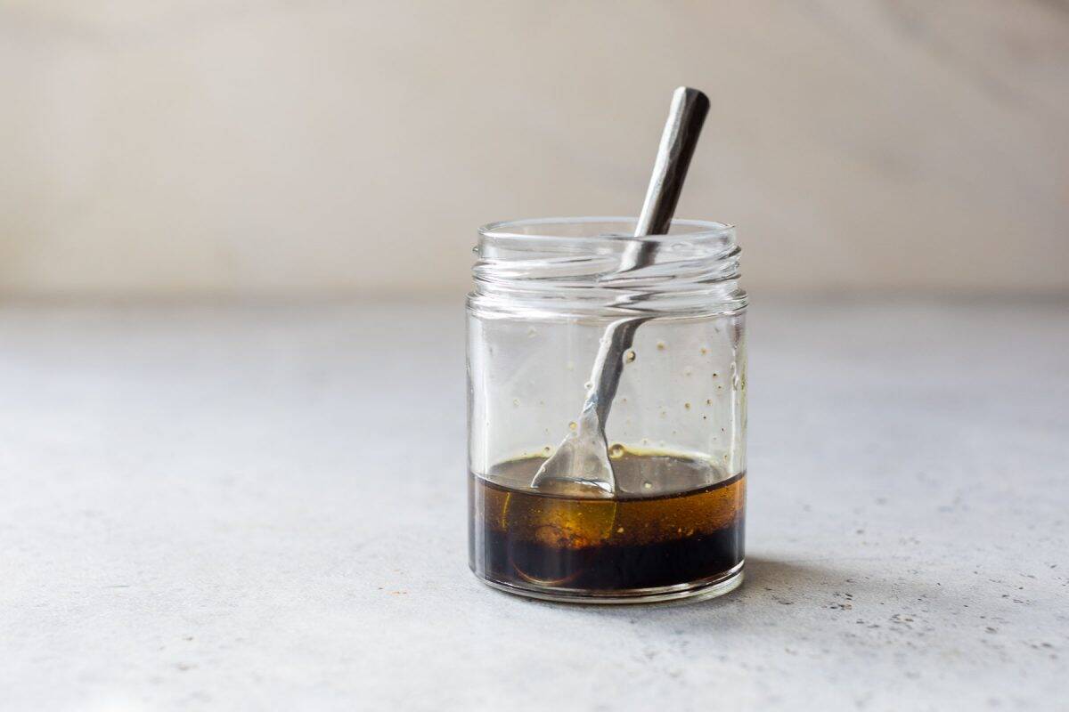 A small glass jar with brown liquid and a metal spoon inside, placed on a light gray surface.
