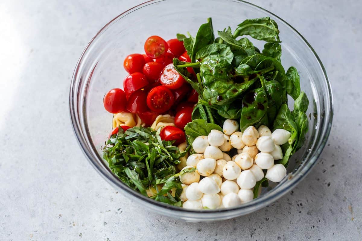 Glass bowl with spinach, cherry tomatoes, mozzarella balls, chopped basil, and tortellini on a light surface.