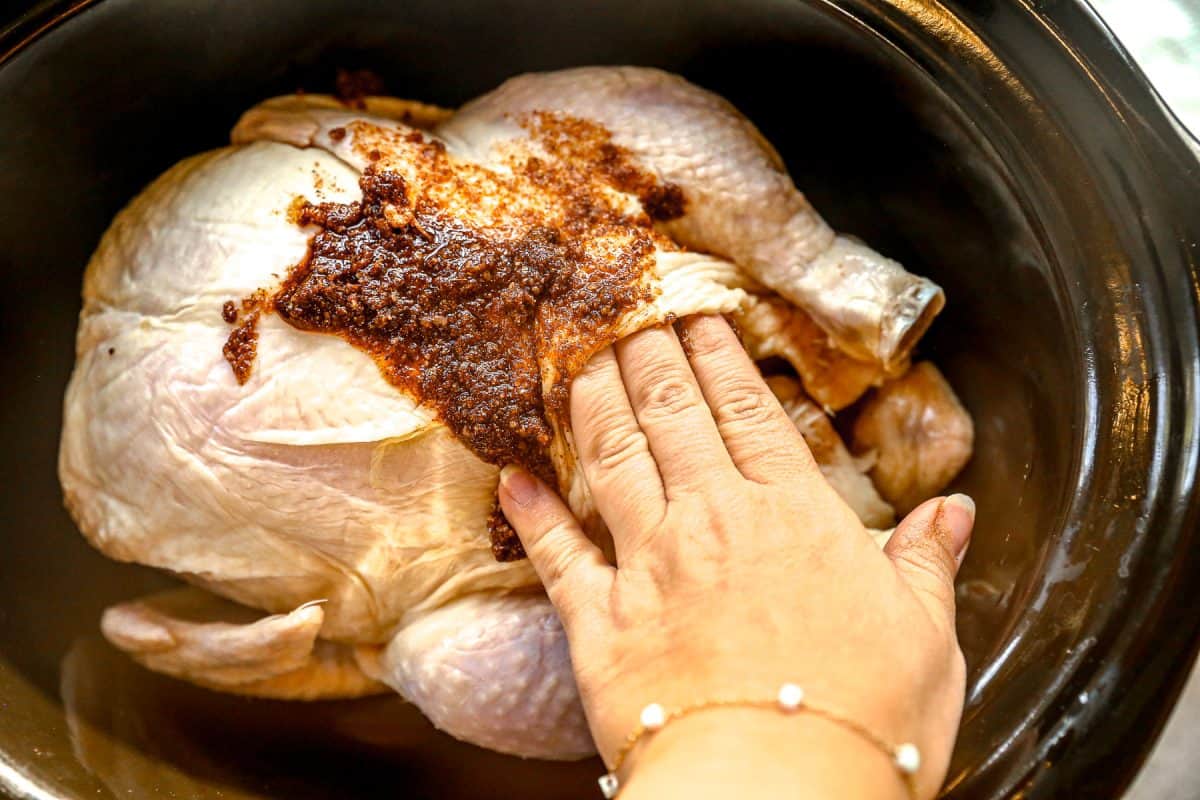 A hand spreads spice rub onto a raw whole chicken inside a black slow cooker.