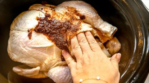 A hand spreads spice rub onto a raw whole chicken inside a black slow cooker.