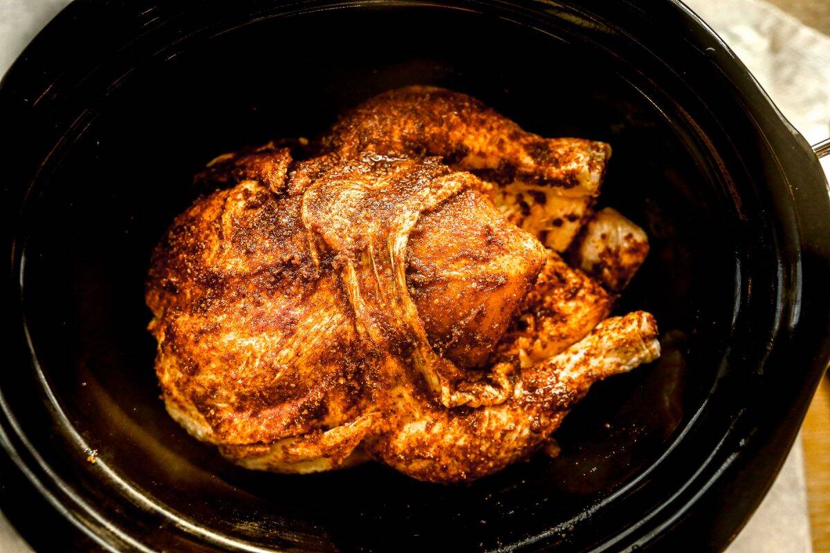 A whole seasoned roasted chicken sits in a black slow cooker, ready to be served.