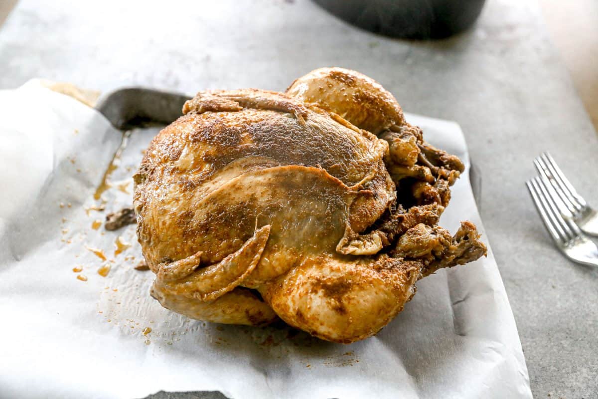 A whole crockpot chicken with seasoning sits on parchment paper beside two forks on a gray surface.