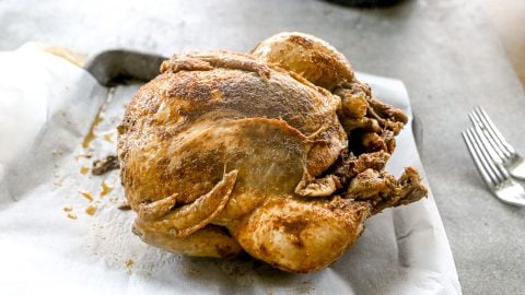 A whole crockpot chicken with seasoning sits on parchment paper beside two forks on a gray surface.