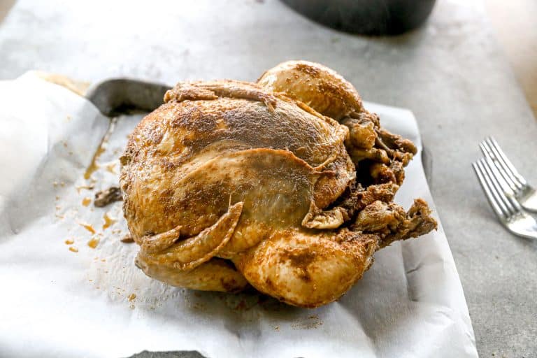 A whole crockpot chicken with seasoning sits on parchment paper beside two forks on a gray surface.