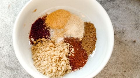 A white bowl with piles of various spices, including brown, red, and beige powders, on a gray surface.