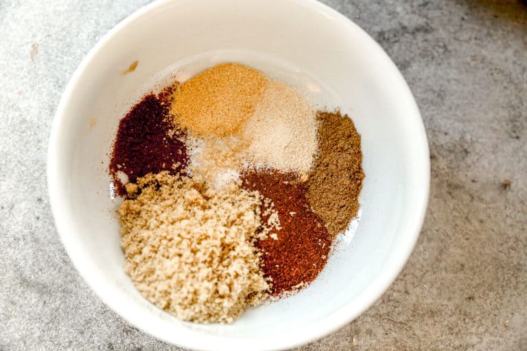A white bowl with piles of various spices, including brown, red, and beige powders, on a gray surface.