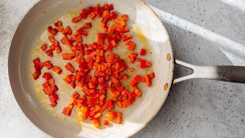 Diced red bell peppers sautéing in a light-colored pan on a gray countertop with sunlight streaming in.