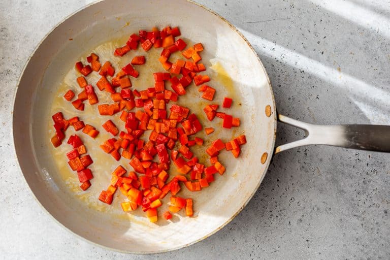 Diced red bell peppers sautéing in a light-colored pan on a gray countertop with sunlight streaming in.