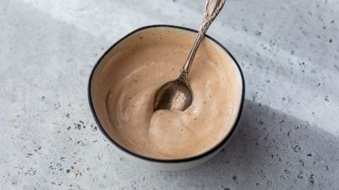 A small bowl of creamy BBQ yogurt sauce with a spoon in it, on a light textured surface.