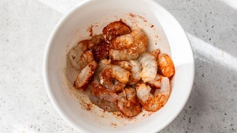 Raw shrimp in a white bowl, seasoned with spices and herbs, on a light textured surface.