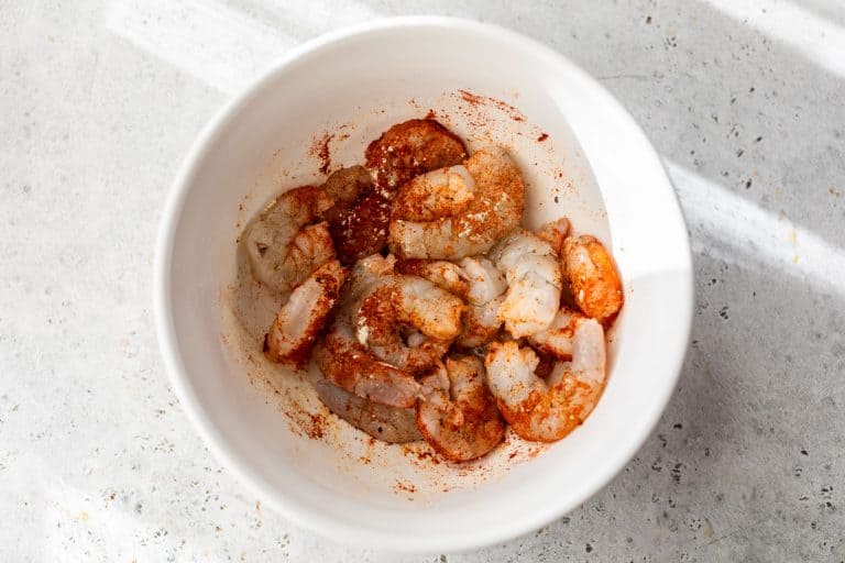 Raw shrimp in a white bowl, seasoned with spices and herbs, on a light textured surface.