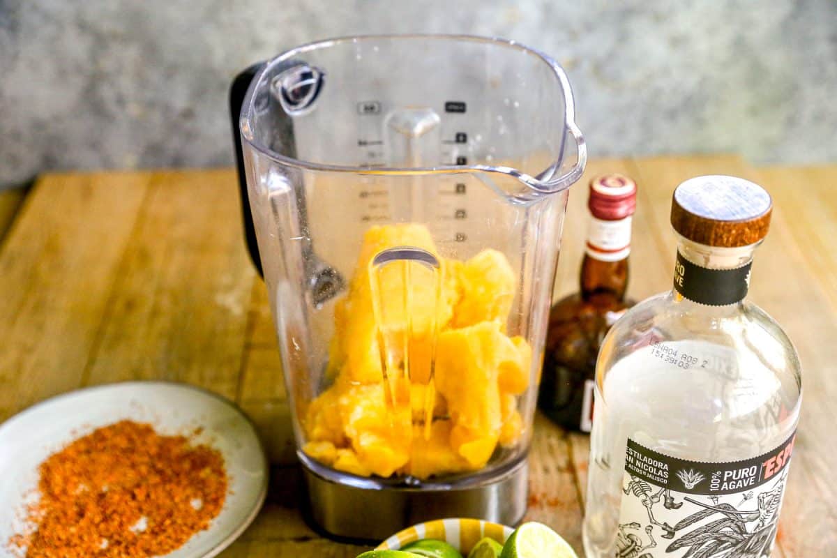 Blender with mango chunks, surrounded by a plate of chili powder, limes, bitters, and a bottle of tequila.