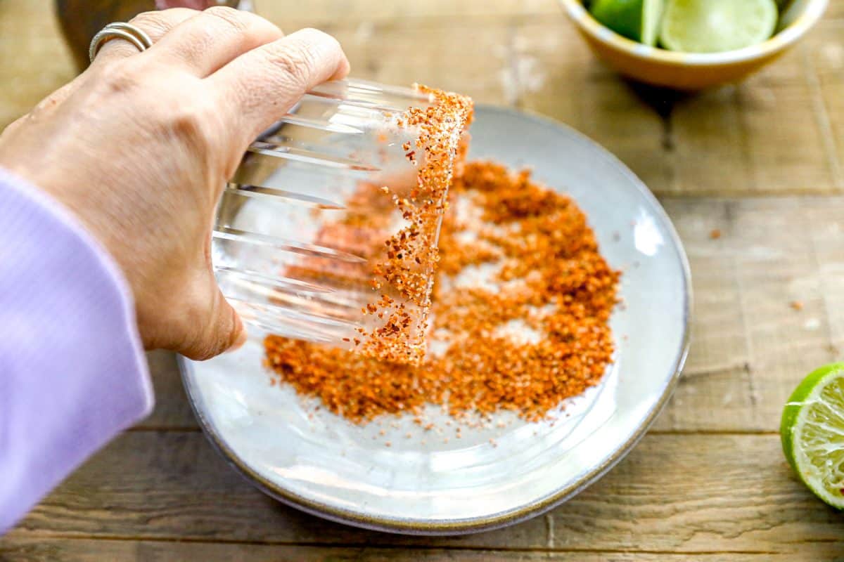 A hand dips a glass rim into a plate of chili seasoning, with lime wedges nearby on a wooden surface.