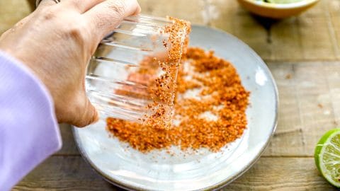 A hand dips a glass rim into a plate of chili seasoning, with lime wedges nearby on a wooden surface.
