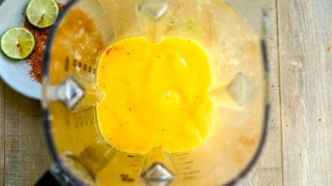 Top view of a blender with mango margarita inside; lime slices and chili powder on a plate beside it.