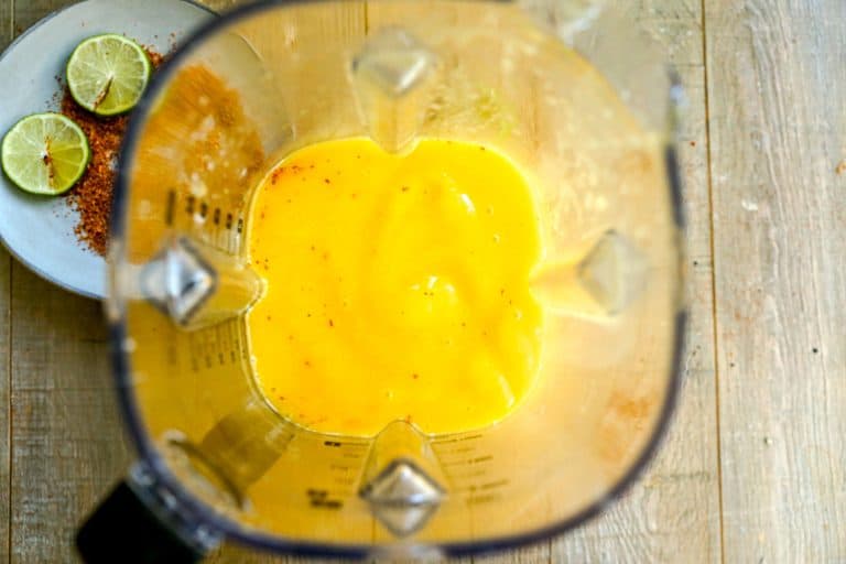 Top view of a blender with mango margarita inside; lime slices and chili powder on a plate beside it.