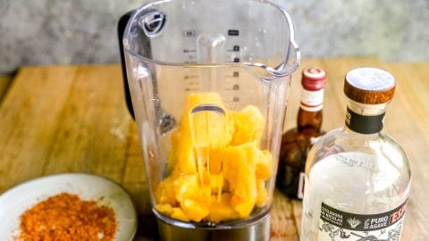 Blender with mango chunks, surrounded by a plate of chili powder, limes, bitters, and a bottle of tequila.