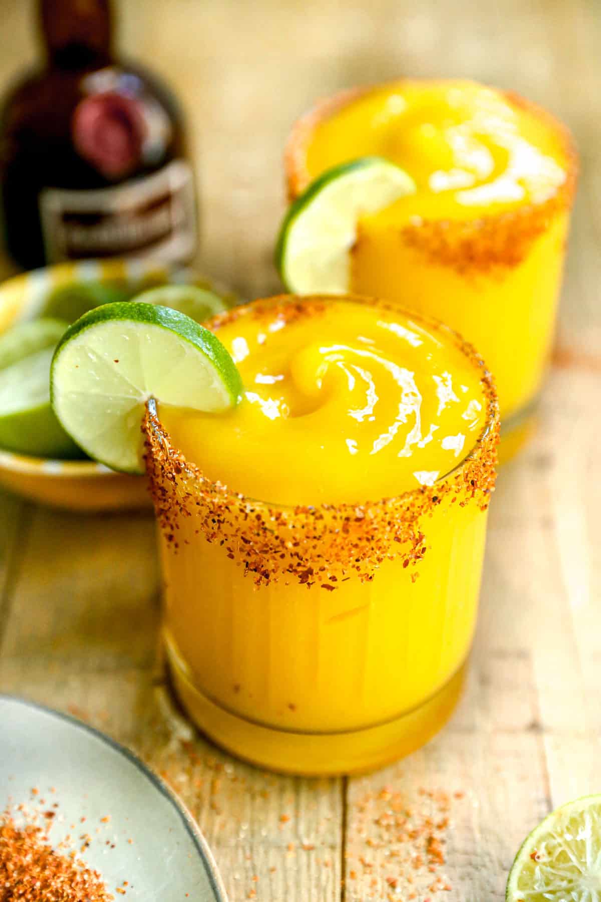 Two glasses of frozen mango margarita with chili powder rims and lime wedges, on a wooden table.