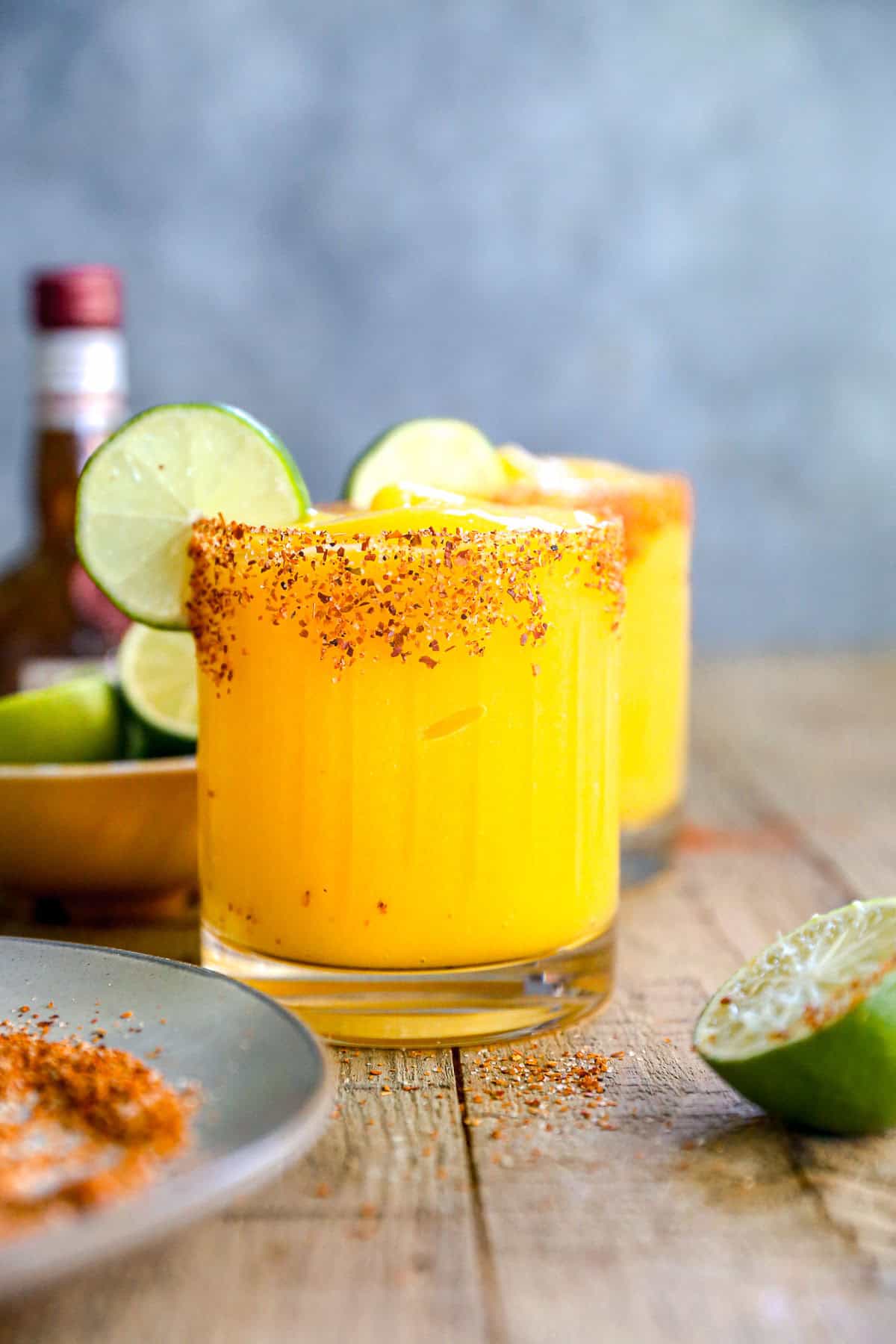A mango margarita with a chili-rimmed glass, garnished with lime slices, sits on a wooden table.
