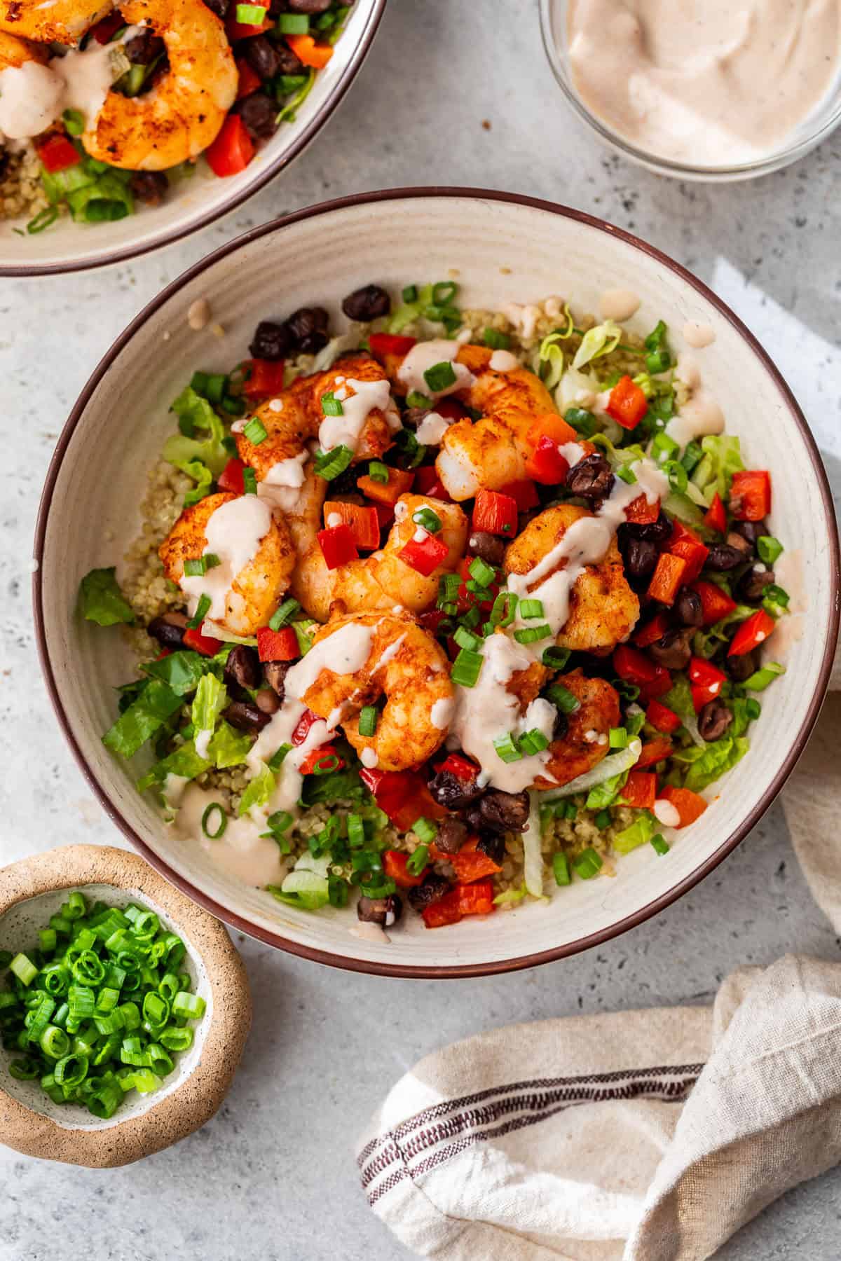 A bowl of seasoned shrimp, black beans, quinoa, bell peppers, and green onions drizzled with creamy sauce.