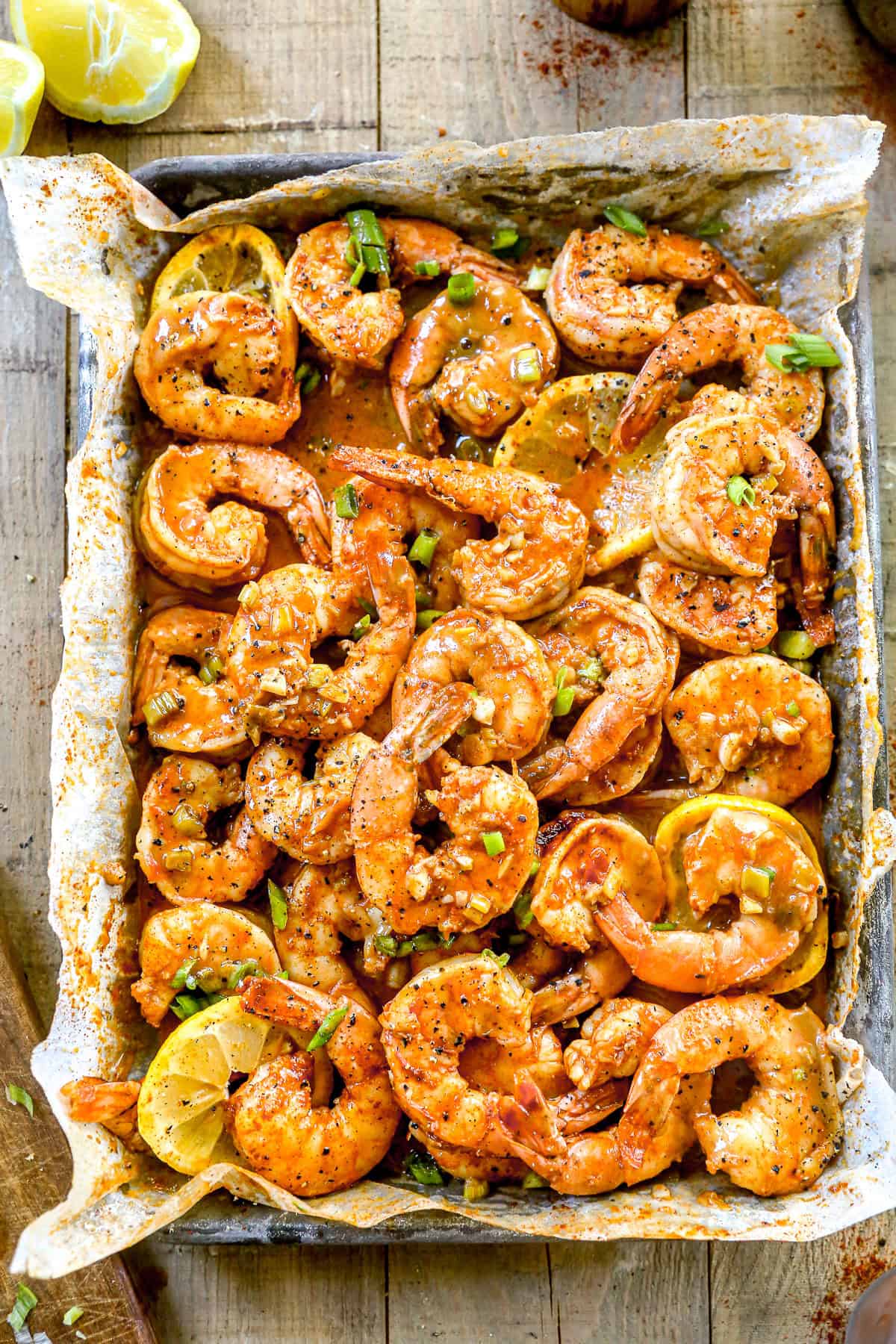 BBQ shrimp with spices and lemon slices on parchment paper, garnished with chopped green onions.