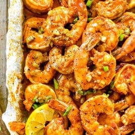Cooked, seasoned BBQ shrimp garnished with chopped green onions and lemon slices on a parchment-lined tray.