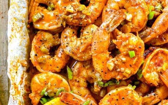 Cooked, seasoned BBQ shrimp garnished with chopped green onions and lemon slices on a parchment-lined tray.