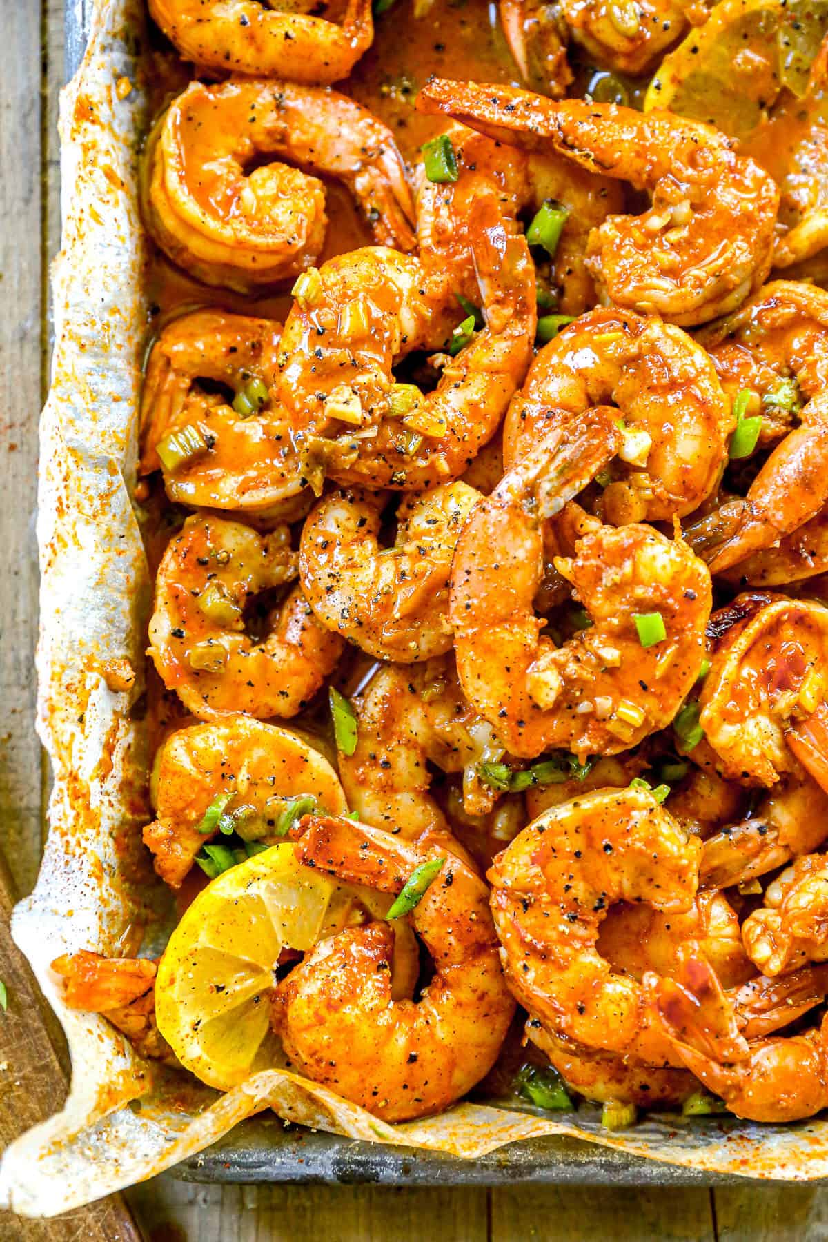 Cooked, seasoned BBQ shrimp garnished with chopped green onions and lemon slices on a parchment-lined tray.