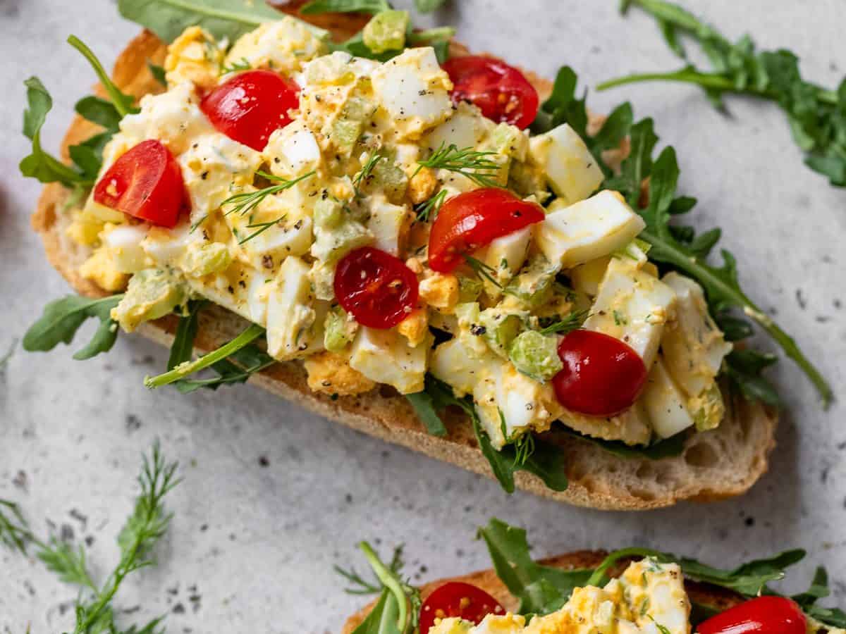 Two slices of bread topped with arugula, healthy egg salad, chopped tomatoes, and fresh herbs on a light surface.