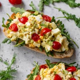 Two slices of bread topped with arugula, healthy egg salad, chopped tomatoes, and fresh herbs on a light surface.