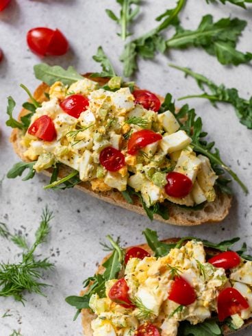 Two slices of bread topped with arugula, healthy egg salad, chopped tomatoes, and fresh herbs on a light surface.