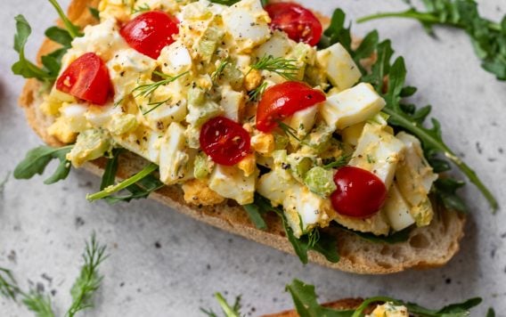 Two slices of bread topped with arugula, healthy egg salad, chopped tomatoes, and fresh herbs on a light surface.