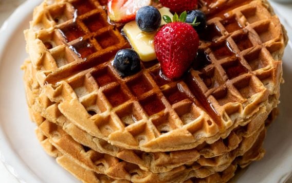 Stack of waffles topped with butter, syrup, strawberries, and blueberries on a white plate.