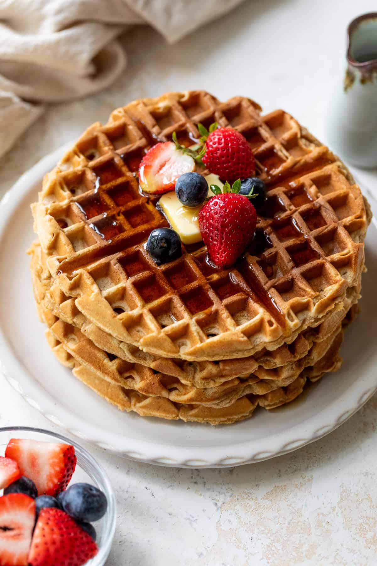 Stack of waffles topped with butter, syrup, strawberries, and blueberries on a white plate.