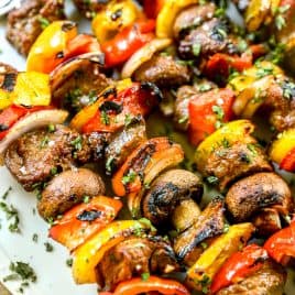 Grilled steak kabobs with peppers, onions, and mushrooms on a white plate, sprinkled with herbs.